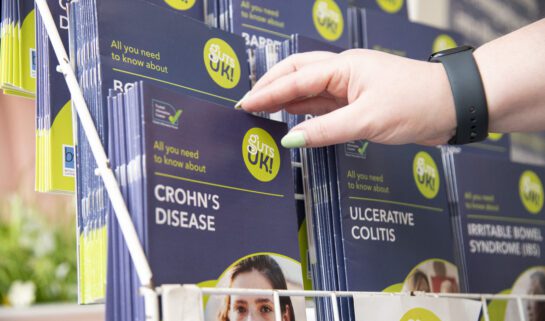 Guts UK's patient information leaflets are displayed in a stand. A hand reaches out to pick up the Crohn's disease leaflet.