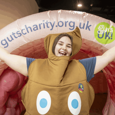 Frankie, a member of the Guts UK team, stands in front of Colin the inflatable colon. Frankie is wearing a poo costume and has her arms up in the air as she smiles at the camera.