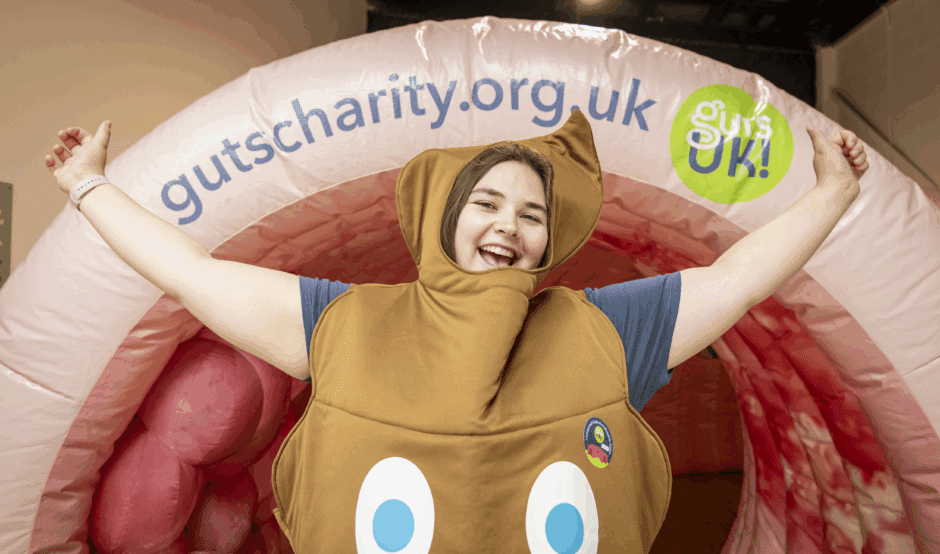 Frankie, a member of the Guts UK team, stands in front of Colin the inflatable colon. Frankie is wearing a poo costume and has her arms up in the air as she smiles at the camera.