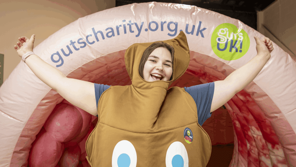 Frankie, a member of the Guts UK team, stands in front of Colin the inflatable colon. Frankie is wearing a poo costume and has her arms up in the air as she smiles at the camera.