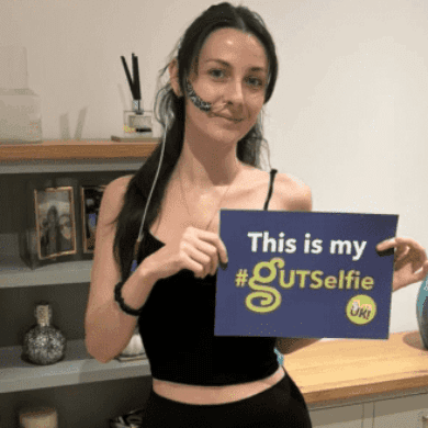 Jessie has long black hair, in a side ponytail over her shoulder. She is wearing a black vest top and leggings, and socks. She has a feeding tube going into her nose, with navy tape holding it to her left cheek. She is holding a #GUTSelfie board and smiles at the camera, in front of a grey bookcase and cabinet.