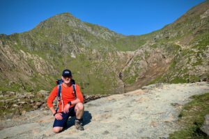 Shawn is wearing a red shirt and kneels on a rocky mountain path, smiling under a clear blue sky. Majestic green mountains tower in the background.