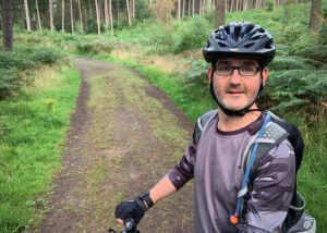 Shawn is wearing a helmet, glasses, and gloves stands with a bike on a forest path. He appears relaxed, surrounded by lush greenery and tall trees.