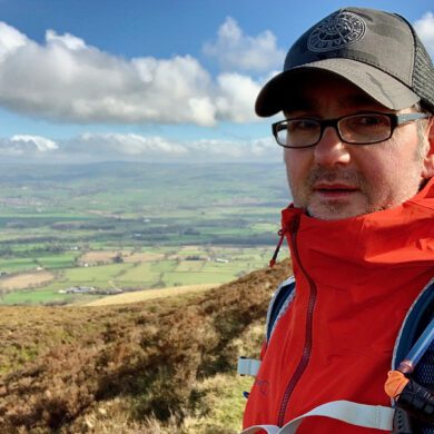 Shawn, wearing glasses, a cap, and a red jacket, stands on a grassy hillside with rolling countryside and a cloudy sky in the background.