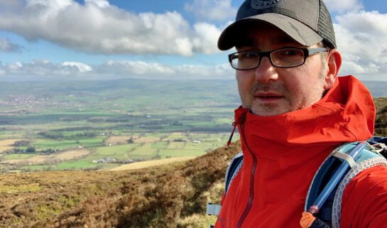 Shawn, wearing glasses, a cap, and a red jacket, stands on a grassy hillside with rolling countryside and a cloudy sky in the background.