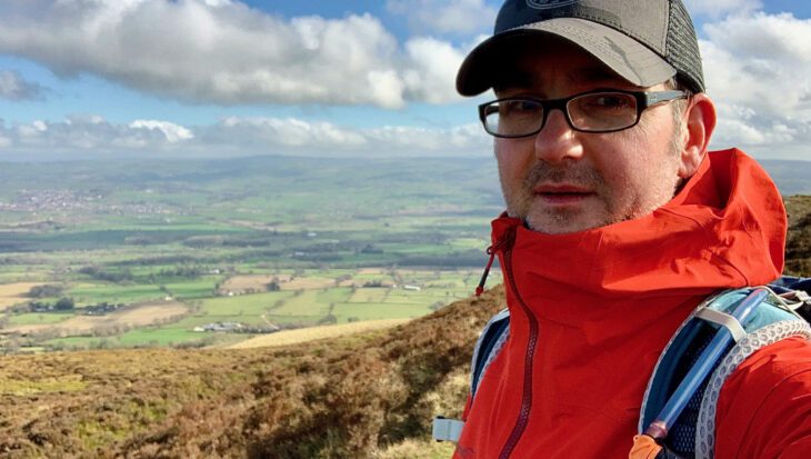 Shawn, wearing glasses, a cap, and a red jacket, stands on a grassy hillside with rolling countryside and a cloudy sky in the background.
