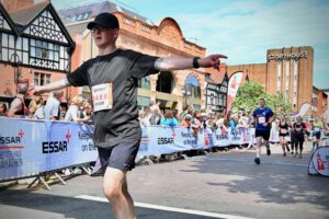 Shawn is seen finishing a race, arms outstretched, in front of cheering crowd. Bright day, historic buildings in background, joyful atmosphere.