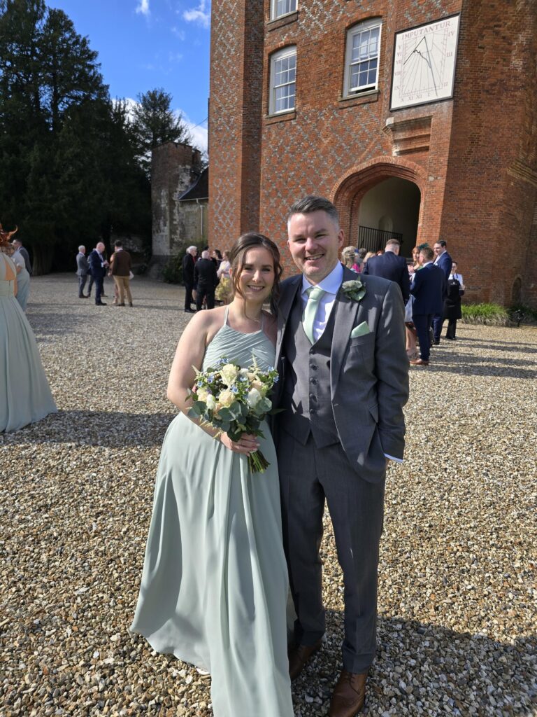 Stephen and his wife are at a wedding together. Stephen wears a three-piece dark grey suit with a pale green tie and pocket square and his wife wears a matching, pale green floaty dress. She is holding a floral bouquet which has cream and pale yellow flowers and greenery. They smile at the camera.