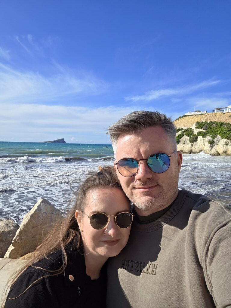 Stephen and his wife are taking a selfie in front of lush, blue sea. The sky is a bright blue and has wisps of clouds.