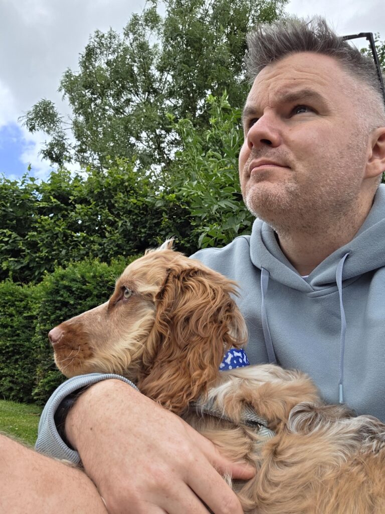 Stephen holds his brown, cocker spaniel puppy, Percy in his arm and is taking a selfie. They are both looking to the left. Stephen wears a light blue hoodie and has his sunglasses rested on his head. He has short brown hair.