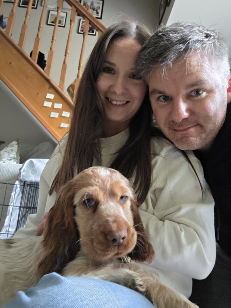 Stephen, his wife and their brown cocker spaniel puppy, Percy, are smiling towards the camera. Stephen has short brown hair and leans on his wife's shoulder, taking the photo. His wife has long, straight brunette hair and wears a cream sweatshirt.
