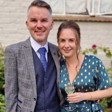 Stephen and his wife are dressed up for a celebration. Stephen has short brown hair, wears a navy waistcoat and trousers, with a grey checked jacket. He also wears brown shoes and a blue tie. His wife wears a teal wrap dress, with gold flecks on, and golden sandals. She has light brown tied up, with a curl down the side of her face. They are both smiling, and Stephen has his arm wrapped around her.