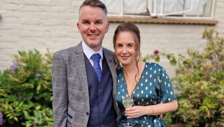 Stephen and his wife are dressed up for a celebration. Stephen has short brown hair, wears a navy waistcoat and trousers, with a grey checked jacket. He also wears brown shoes and a blue tie. His wife wears a teal wrap dress, with gold flecks on, and golden sandals. She has light brown tied up, with a curl down the side of her face. They are both smiling, and Stephen has his arm wrapped around her.