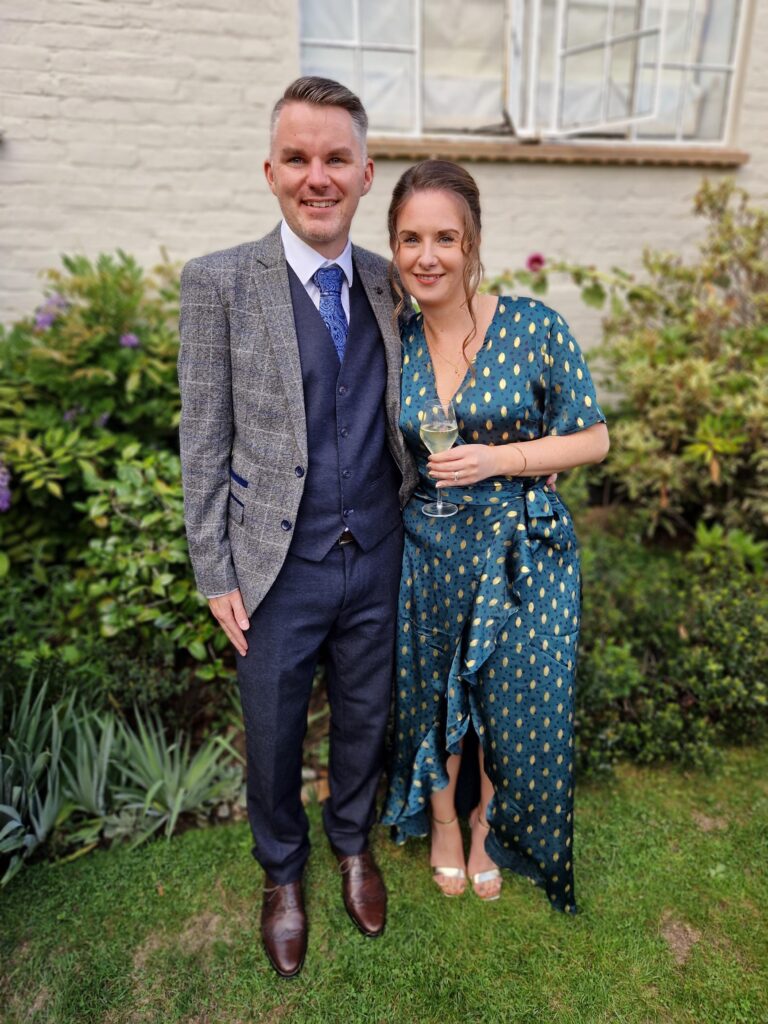 Stephen and his wife are dressed up for a celebration. Stephen has short brown hair, wears a navy waistcoat and trousers, with a grey checked jacket. He also wears brown shoes and a blue tie. His wife wears a teal wrap dress, with gold flecks on, and golden sandals. She has light brown hair tied up, with a curl down the side of her face. They are both smiling, and Stephen has his arm wrapped around her.