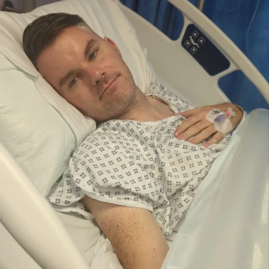 Stephen is in a hospital bed, wearing a hospital gown. He has a cannula in his hand and looks at the camera.