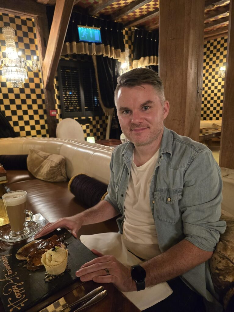 Stephen is sat at a restaurant with a coffee and sausages and mash in front of him. There is cream and black pattern on the wall in the background and a glass chandelier. He wears a white t-shirt with a light blue denim overshirt, his wedding ring, a black watch and has light brown, short hair. He smiles at the camera.