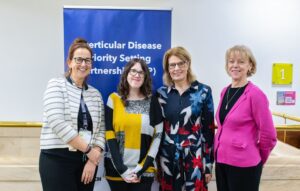 A group of four people stand in a line, smiling at the camera. They are Suzanne (Guts UK), Sam (BRUK), Helen (Guts UK) and Lindsay (BRUK). There is a blue pop up banner behind them that reads: Diverticular Disease Priority Setting Partnership (PSP).