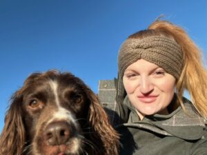 Rachel is smiling, wearing a beige headband with her brown Spaniel. They are outdoors under a clear blue sky.
