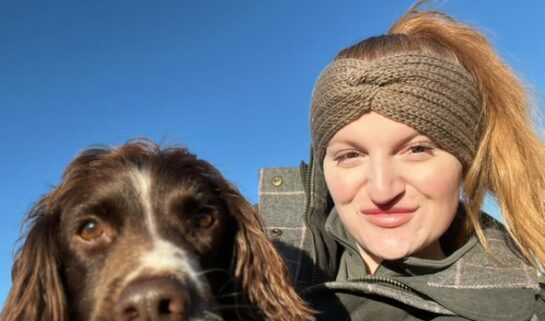 Rachel is smiling, wearing a beige headband with her brown Spaniel. They are outdoors under a clear blue sky.