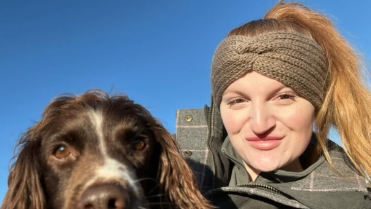Rachel is smiling, wearing a beige headband with her brown Spaniel. They are outdoors under a clear blue sky.