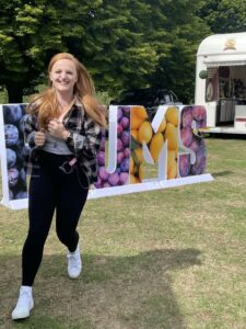 Rachel is running happily on the grass. In the background, large colorful letters spell out 'PLUMS' near a food truck.