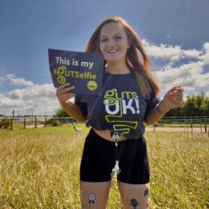 Rachel is standing in a sunny field, holding a #GUTSelfie sign that reads: "This is my #GUTSelfie." She is wearing a navy blue Guts UK t-shirt and black shorts. The #GUTSelfie temporary tattoos are seen on her thighs.