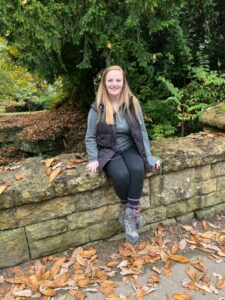 Rachel is sitting on a stone wall in a park, surrounded by autumn leaves and lush greenery. She wears a vest, sweater, and hiking boots and is smiling warmly.