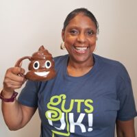 Jackie is smiling at the camera and wears a Guts UK t-shirt. She is holding a smiling poop emoji mug in her hand.
