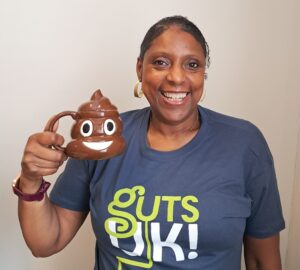 Jackie is smiling at the camera and wears a Guts UK t-shirt. She is holding a smiling poop emoji mug in her hand.