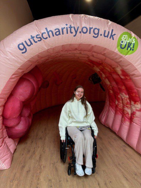 Guts UK supporter, Lucy, sat in her wheelchair in front of colon the giant colon, smiling at the camera. 
