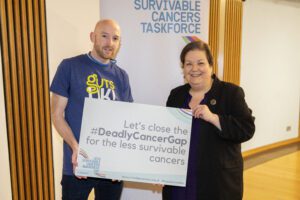 Ross, a family advocate, is pictured with a member of Scottish Parliament, Jackie Baillie, in Scotland. They are holding a Less Survivable Cancers Taskforce sign and smiling at the camera.