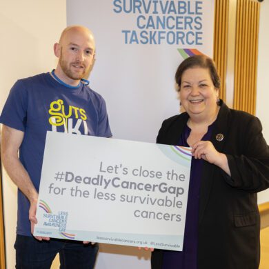 Ross, a family advocate, is pictured with a member of Scottish Parliament, Jackie Baillie, in Scotland. They are holding a Less Survivable Cancers Taskforce sign and smiling at the camera.