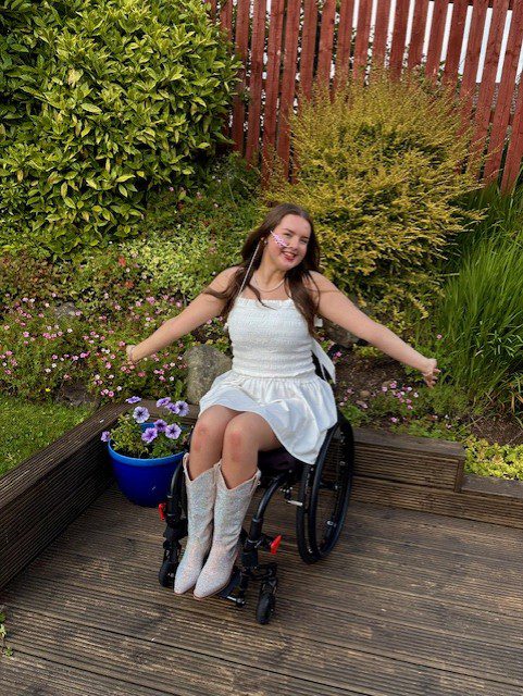 Lucy sat in her wheelchair in a garden, smiling with her arms stretched out joyfully.