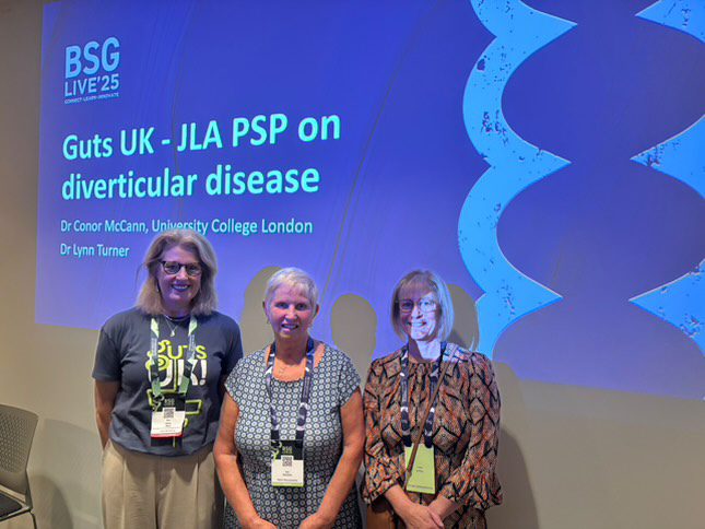 Sue is pictured (centre) next to a Guts UK team member, Helen and another patient, Linda, in front of a screen that reads 'Guts UK- JLA PSP on diverticular disease'.