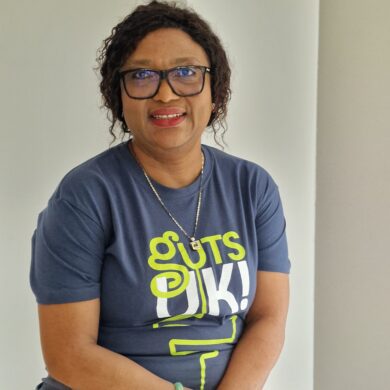 Edith is sat smiling at the camera, with her forearms overlapping, resting her hands on her lap. She wears a navy Guts UK t-shirt