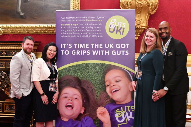 The parents of Grace and the parents of Alia stand together smiling beside a Guts UK banner at St James’s Palace. The banner features photos of Grace and Alia with the words: “It’s time the UK got to grips with guts.”