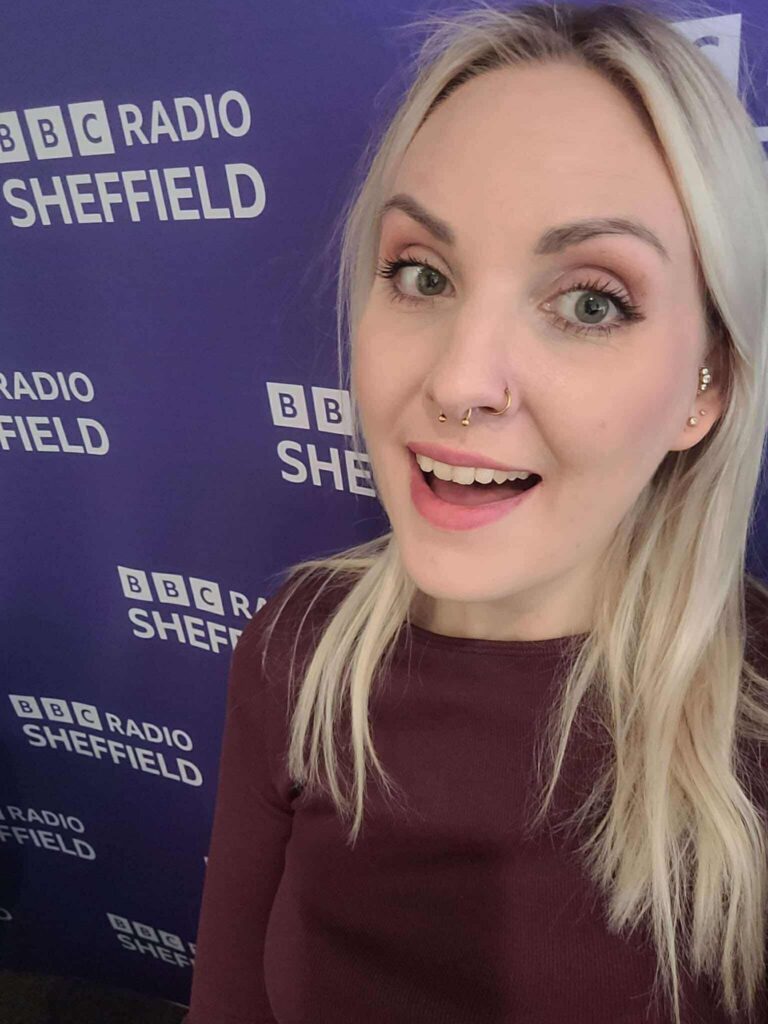 Leanne smiles in a selfie style shot with a purple backdrop with BBC Radio Sheffield branding on it.