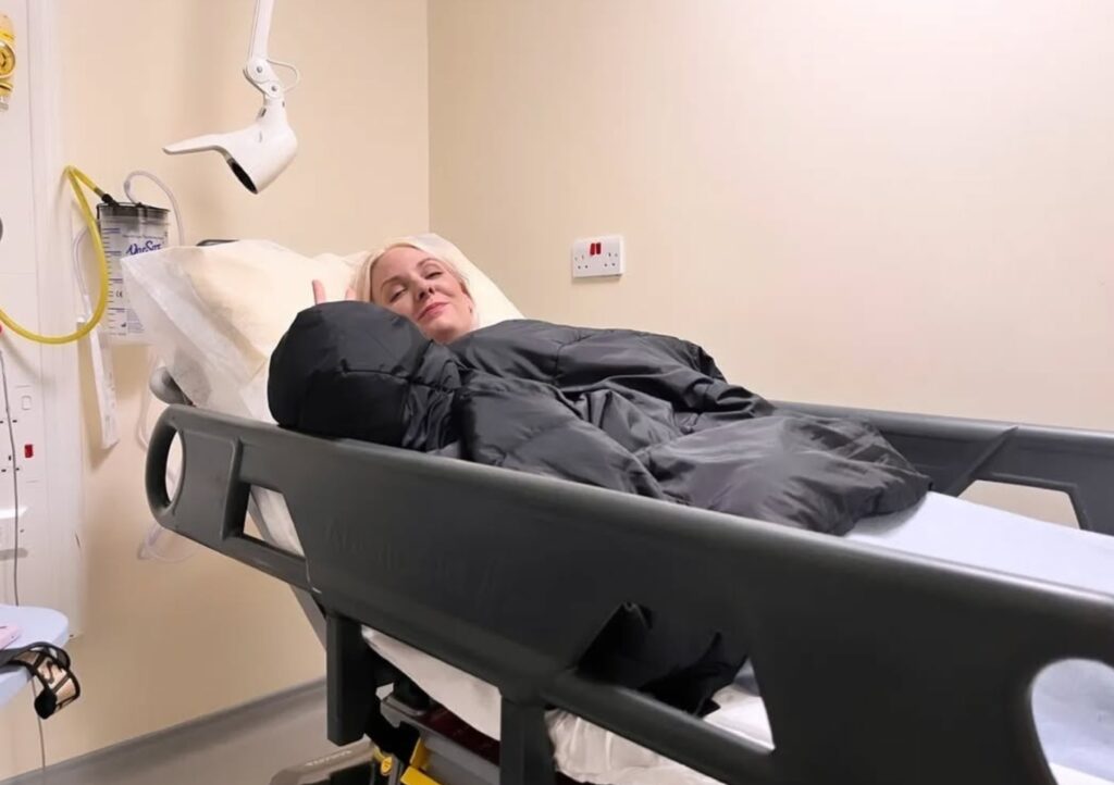 Leanne laid on a hospital trolley, her head slighted tilted to look at the camera, with a thumbs up gesture.