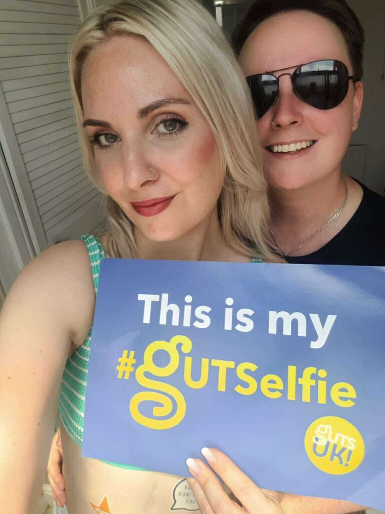 Leanne is smiling in selfie style shot holding a blue card that reads 'This is my #gutselfie.' Her partner stands behind her smiling.