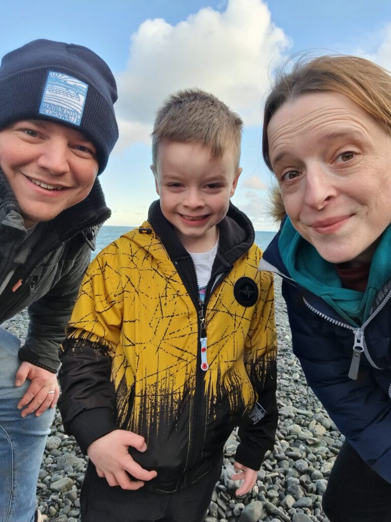 Lisa, Craig and George are smiling at the camera, on a grey, pebbled beach.