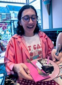 Rachael is wearing a striped pink shirt and holding a plate with chocolate cake with a pink candle. She smiles happily.