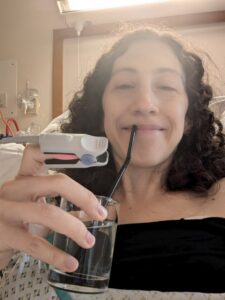 Rachael is in a hospital gown and holding a glass of water with a straw. She wears a pulse oximeter on her finger and is smiling at the camera.