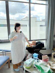Rachael is in a hospital gown and blue slippers. She is smiling at her baby while holding a cup. They are in a bright room with a large window behind.