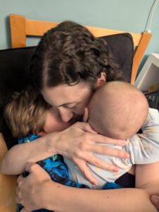 Rachael is sitting on a wooden chair and hugging her two children tightly.