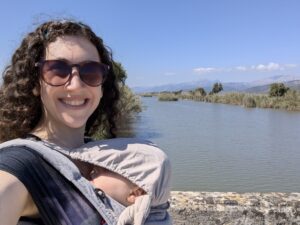 Rachael is smiling and wearing sunglasses while holding her baby in a carrier. She is standing beside a tranquil river with distant mountains under a clear blue sky.