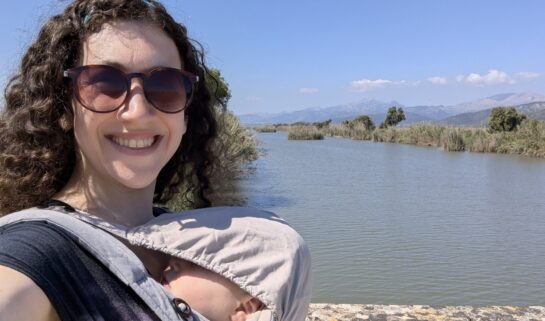 Rachael is smiling and wearing sunglasses while holding her baby in a carrier. She is standing beside a tranquil river with distant mountains under a clear blue sky.