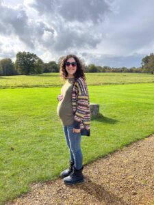 Rachael is standing outdoors on a gravel path next to grass, wearing sunglasses, a patterned cardigan, jeans, and boots, with one hand on their pregnant belly.
