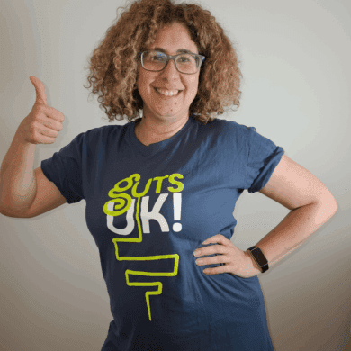 Rawan stands with her hand on her hip, and her left hand help in a 'thumps up' pose. She has light brown, short, curly hair and wears a navy Guts UK t-shirt.