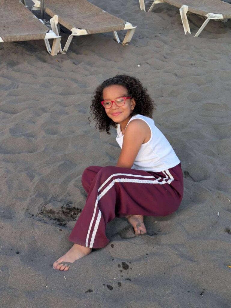 Alia sits on the sand at the beach, smiling at the camera. She is wearing red glasses, a white vest, and maroon trousers with white stripes.