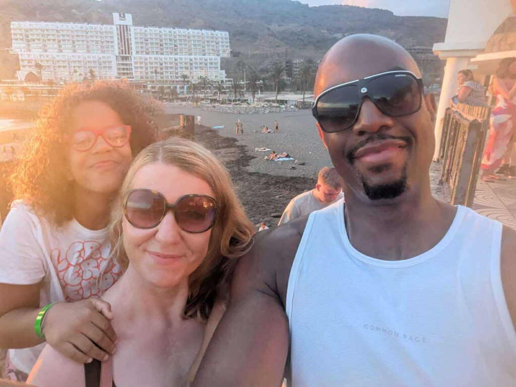 Alia is pictured with her mum and dad on a beach at sunset. They are smiling for a selfie, with the sea and a large hotel building in the background.